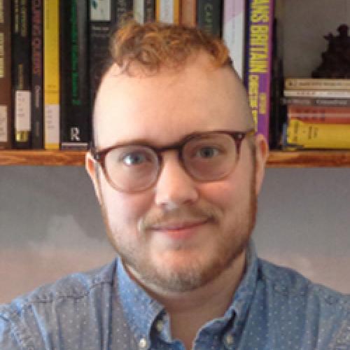 Adrian Kane wears glasses and a blue shirt with a bookcase in the background.