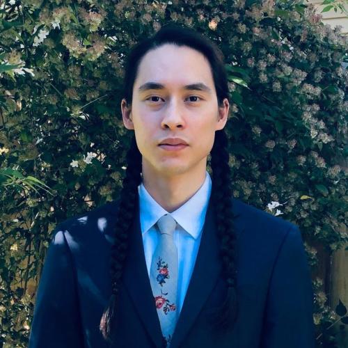 Andreas P. Bassett stands in front of a large shrubbery while wearing a dark jacket blue shirt and tie.