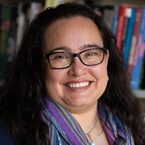 Portrait of Ariana Ochoa Camacho wearing glasses and standing in front of a bookcase.