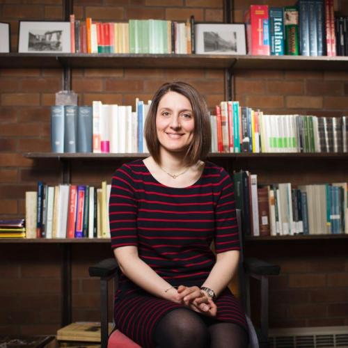 Profile of Beatrice in front of a book case. She has short brown hair and is wearing a sweater while smiling at the camera.