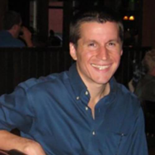 Nathaniel Bond wears a blue collared shirt while sitting in a chair and looking into the camera.