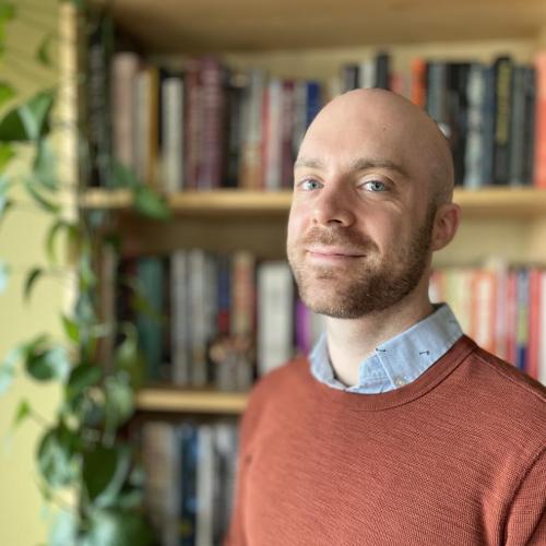 Caleb Knapp wearing blue oxford shirt and burnt orange sweater poses in front of bookshelf for portrait