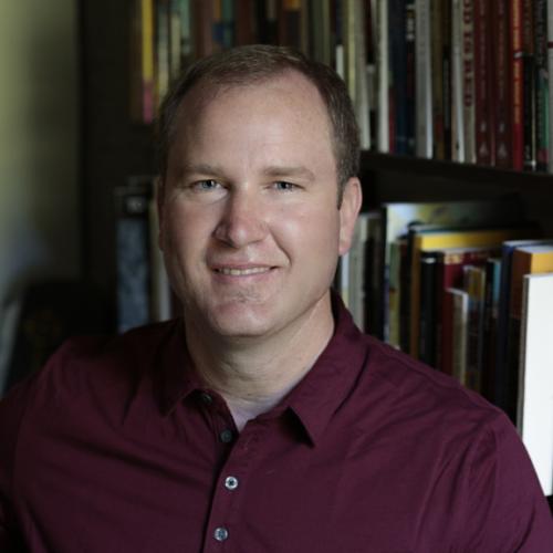 Christopher Teuton sits in front of a bookcase wearing a maroon shirt.