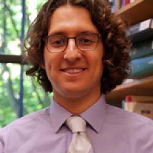 Portrait of Devin Naar wearing a purple shirt and tie and wearing glasses.