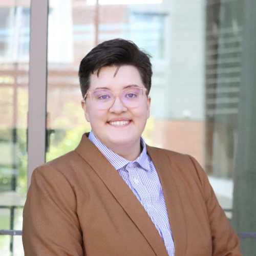 A white butch person with glasses smiling in a light brown blazer and blue and white checkered button-up shirt. 