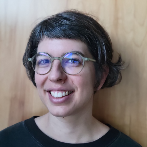 Portrait of Hannah Frydman standing in front of a brown wall wearing glasses.