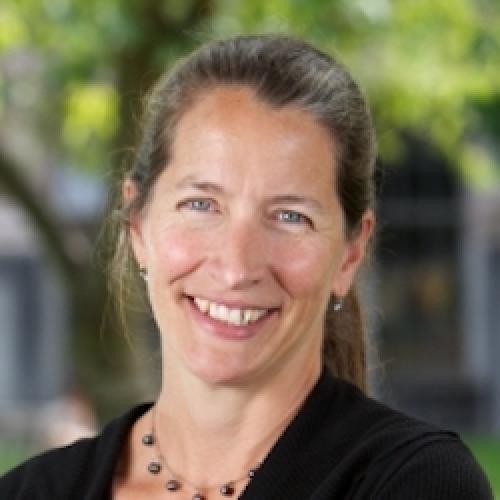 Sara Goering professional photo smiling at the camera, blurred trees in the background, wearing a blue blazer, necklace, and hair pulled back