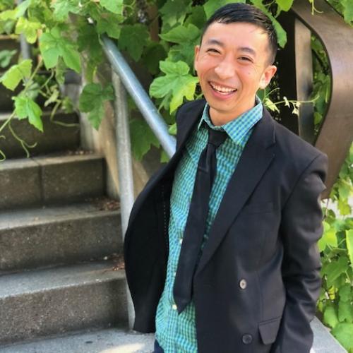 Douglas S. Ishii stands in front of stairs while wearing a blazer, teal collared shirt, and a black tie.