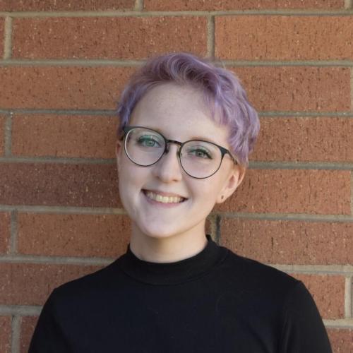 Head and shoulders of a smiling white person with short purple hair and glasses in a black turtleneck against a brick wall background