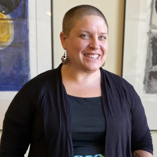 Julie stands in natural light smiling brightly. A thick white woman with a shaved head, she wears a black top and cardigan and large silver and black earrings. Behind her is a cream-colored wall with the framed edges of art pieces hung on each side of her.