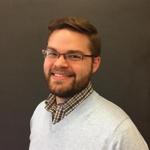 Justin Randolph stands in front of a dark wall while wearing glasses and a white sweater.