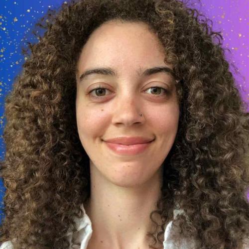 A profile picture of Kelly, a young woman with white skin and big brown curly hair, wearing a white collared shirt and smiling, with a blue to purple gradient background with glitter sparkles.