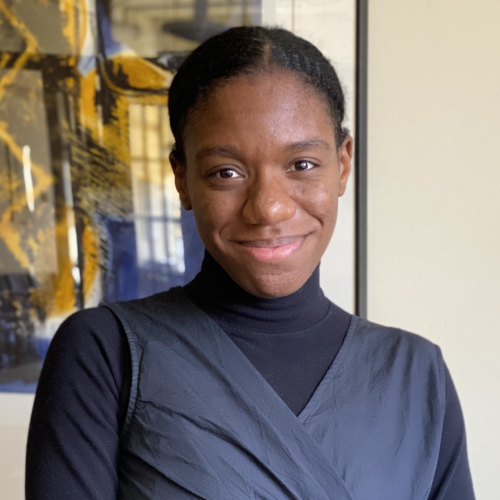 profile of Kalia smiling at the camera, hair back in a bun and in a black turtle neck top with overlapping black dress