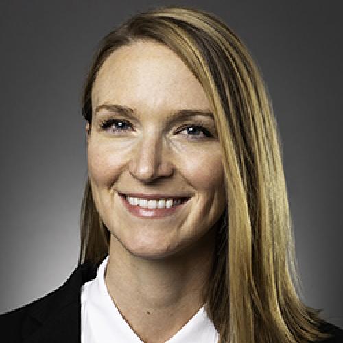 Kristin Dew stands in front of a dark wall wearing a white shirt and dark jacket.