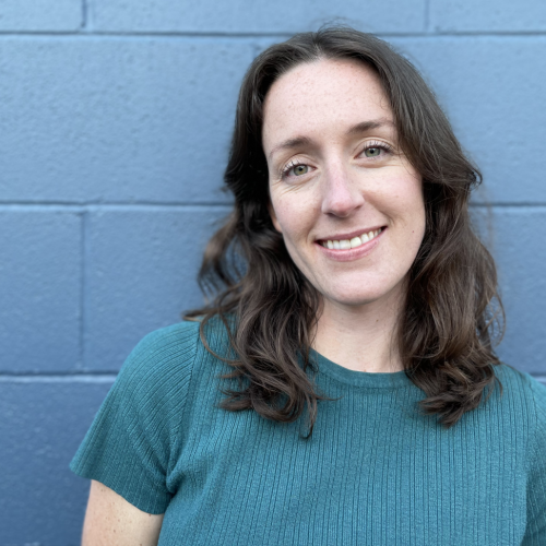 Portrait of Madison, a woman with long brown hair with a blue t-shirt, in front of a blue brick wall. 