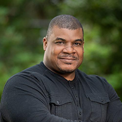 A portrait of Marcus Johnson standing in front of tress wearing a black shirt.