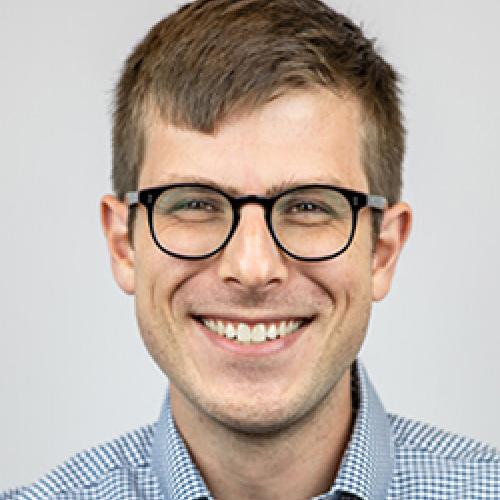 Matthew Bellinger stands in front of a white wall wearing a patterned shirt and glasses.