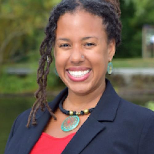 profile of Maya Smith smiling at the camera, hair pulled back with some curls along the left side, wearing a red top under a blue blazer with a necklace