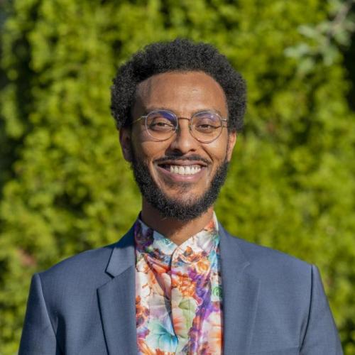 Nathanael in front of a backdrop of green foliage, smiling while wearing a blue blazer and floral shirt.