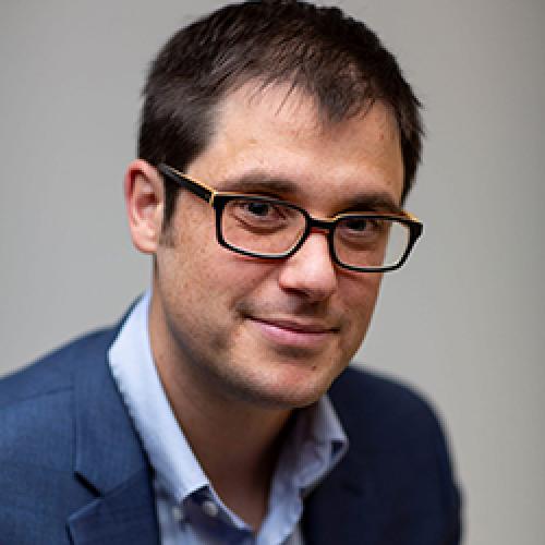 P. Joshua Griffin sits in front of a gray wall wearing glasses, a blue shirt, and a dark jacket.