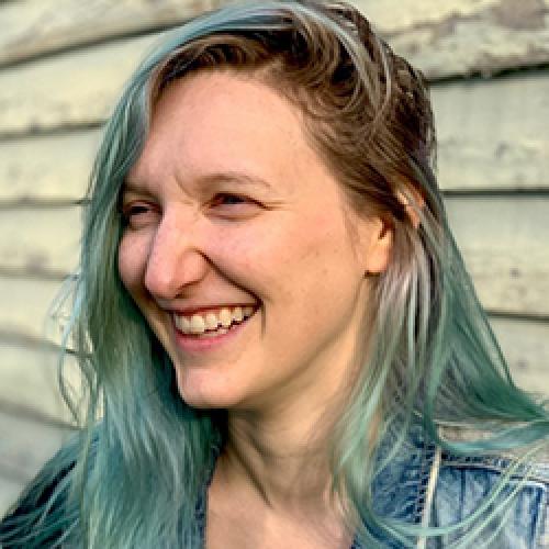 Jacqui Pratt has blue hair and looks to the left and laughs while standing in front of a shiplapped exterior wall.