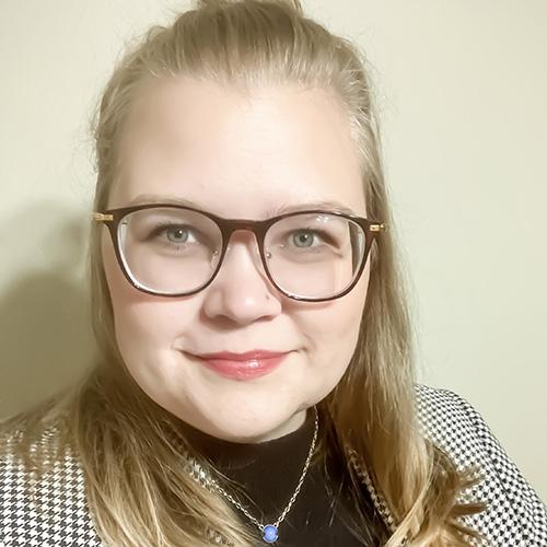 Samantha Thompson stands in front of a beige wall while wearing glasses and a jacket.