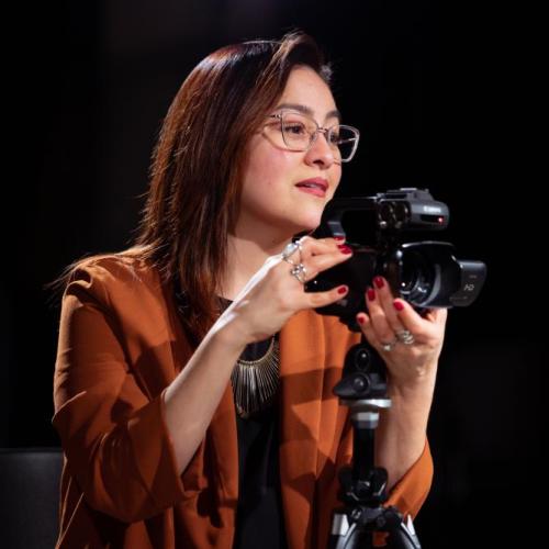 Sonia De La Cruz looks to the right while standing behind a camera on a tripod.