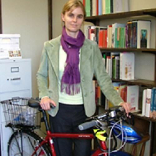 Susan Gaylard stands behind a red bicycle and in front of a bookcase wearing a light-green jacket and purple sweater.