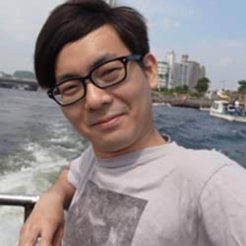 Yuta Kaminishi stands in front of a body of water wearing glasses and a grey shirt.