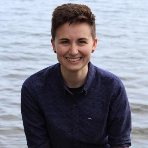 Profile picture of Sarah Brucia Breitenfeld: a person with short brown hair in a blue button up sitting in front of a lake.