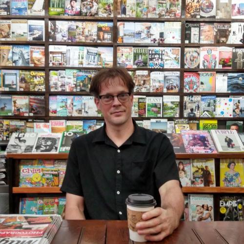 Image of Aaron Carpenter, male, sitting at a wooden table, holding a coffee cup. Magazines are displayed on shelves behind him.