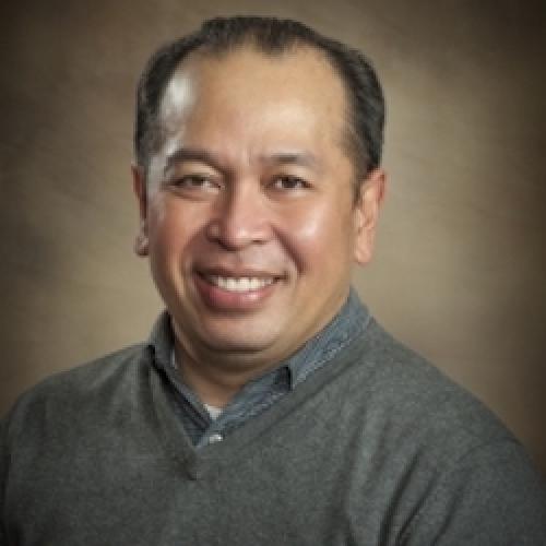 professional photo of Rick Bonus smiling at the camera in a blue shirt and a brown background