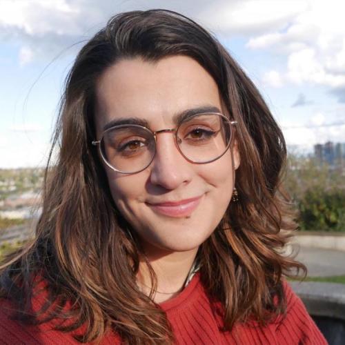 Close-up photo of a smiling woman with brown hair wearing round glasses and a red sweater. Part of the Seattle skyline is in view behind her.