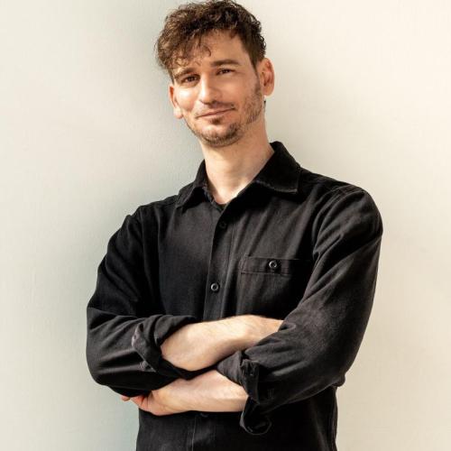 James Pierce stands in front of a white wall with his arms crossed while wearing a black collared shirt and black trousers.