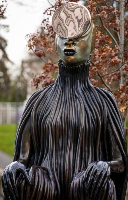 bronze statute of an African Women with an obelisk over her eyes and forehead, a garmetn in ribbons running down her seated body, hands on her knees