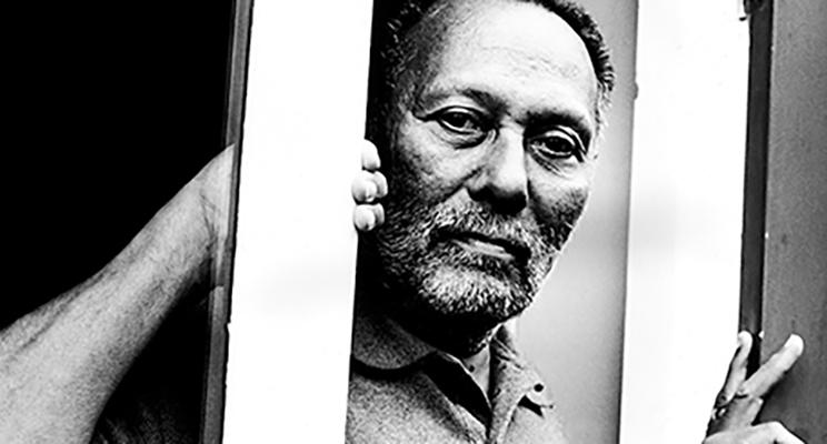 Black and white image of Stuart Hall looking at the camera from behind a pole.