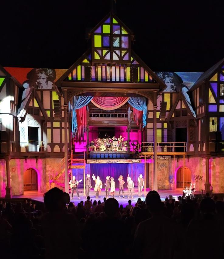 Photo of people on stage with the audience darkened in the foreground and a lit exterior of buildings in the background.