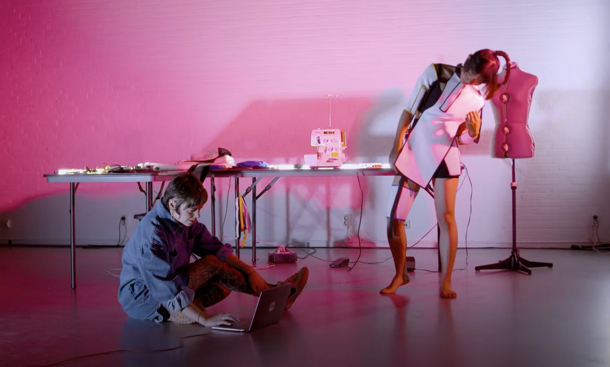 In a magenta lit room one woman sits on the ground with a laptop and the other stands while wearing a white futuristic garment.