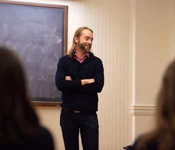  Joel Alden Schlosser stands with arms crossed in front of a chalkboard.