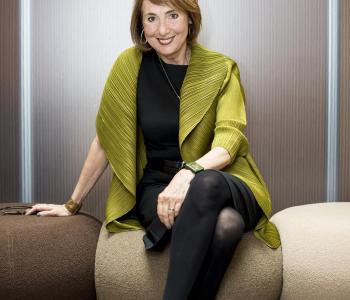 Cathy Davidson smiling at the camera and sitting on a sofa with a green overcoat and black business casual outfit.