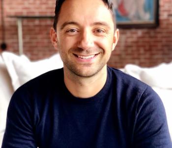 Portrait of Cristian Capotescu wearing a dark blue long sleeved shirt and sitting on a bed in front of a brick wall.