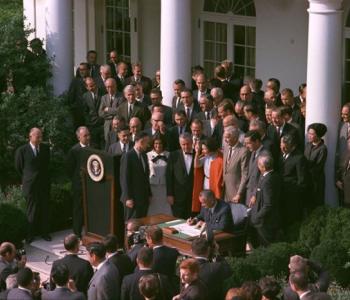 President Lyndon Johnson sits at a desk and signs a document while people in suits stand around him.