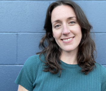 Portrait of Madison Snider: curled, shoulder-length brown hair, blue eyes, smiling at the camera. Blue brick background and wearing a blue t-shirt.