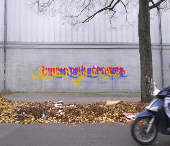 Yellow, blue, and red writing on a grey wall behind a gutter piled high with autumn leaves and street litter. On the right is a tall bare tree and an old Berliner biker zipping into the frame.