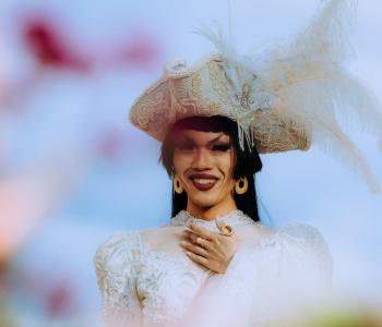 Asian American women in a white hat and dress smiling against a blue sky. Out of focus flowers in the foreground.