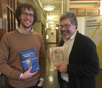 Jason Groves and Jose Alaniz each hold their books while standing in a hallway in front of a sign for the Simpson Center.