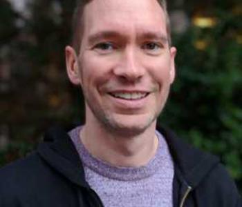 A close-up image of Brendan McElmeel smiling and wearing a purple shirt with a black jacket.