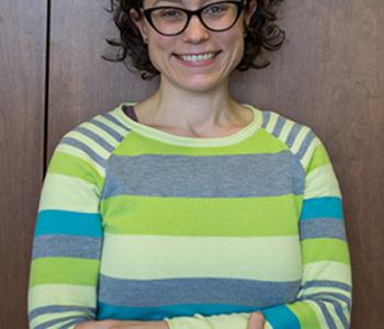 Annie Dwyer stands in front of a wood-paneled wall with arms crossed, wearing a striped green sweater.