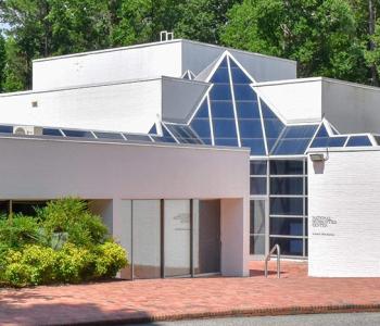 entrance to the national humanities center, which is a white, modern building with angled ceiling windows and a brick courtyard entrance surrounded by green trees and bushes