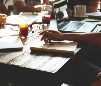 People sit around a wooden table that has notebooks, laptops, and coffee on it.